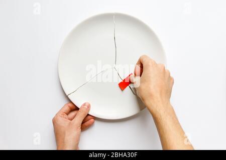 Top view of man hands holding a broken white plate, glues parts with red tape. Metaphor for divorce, relationships, friendships, crack in marriage. Is Stock Photo