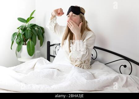 a European woman with blonde hair is sitting on a white bed, she just woke up in the morning. she is dressed in light clothing and a sleep mask. she d Stock Photo