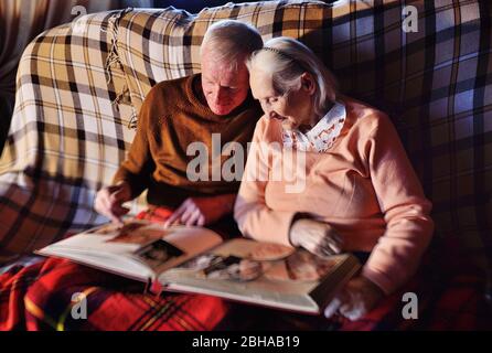 elderly married couple-a man and a woman in a cozy house sitting on the sofa and wrapped in a warm plaid plaid hug each other and smile while viewing Stock Photo