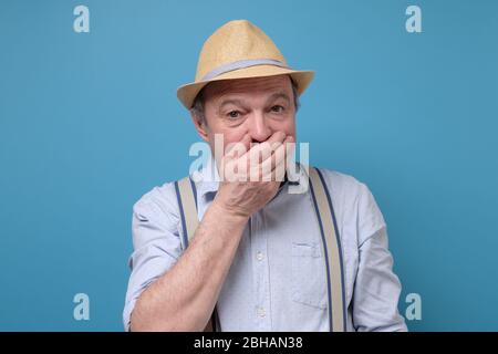 Stunned elderly man in summer hat covers mouth with hands, being surprised Stock Photo