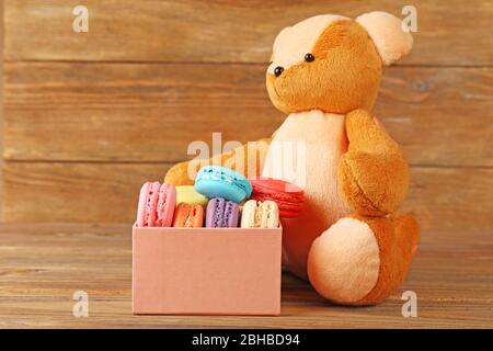 Tasty colorful macaroons in present box near teddy bear and female hand on rustic wooden planks background Stock Photo