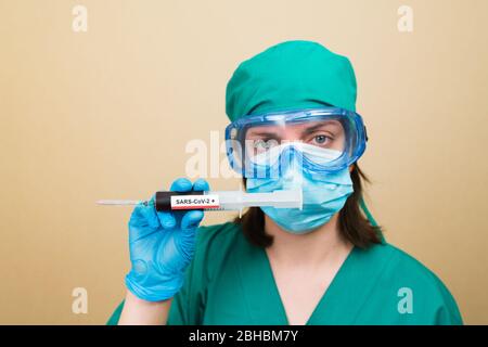 Female doctor in green suit and blue gloves holds test tube with positive COVID-19 blood on the yellow background. Medical concept. Selective focus.