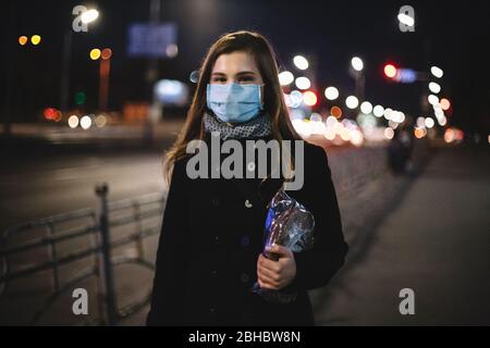 Portrait of young happy woman wearing face medical mask carrying bread while walking on city street at night