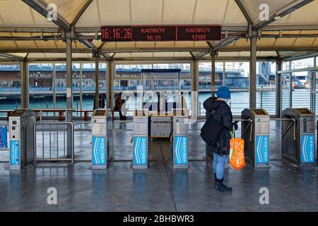 Woman with skullcap and face mask holding shopping bag waiting for Eminonu - Kadikoy ferry in front of toll gates. Galata Bridge on the background. Stock Photo