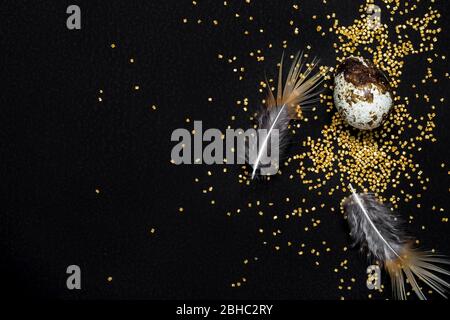 Moody easter background. Quail eggs, feathers and golden sugar sprinkles on black background. Happy easter. Stock Photo