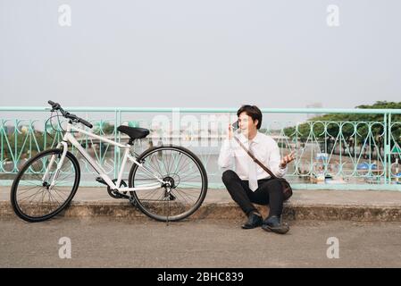 Young asian man talking by mobile phone while sitting on the ground near the bike, outdoors. Wearing stylish clothes. Stock Photo