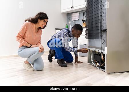 Young Repairman Repairing Refrigerator In Front Of Beautiful Woman Stock Photo