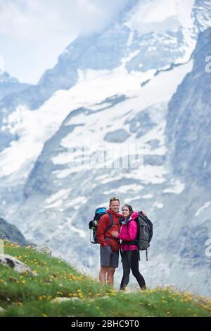 Portrait of couple of tourists travelling in Swiss Alps, man and woman standing on green meadow full of flowers on a breathtaking background with amazingly beautiful rocky mountains Stock Photo