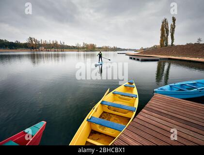 Man on the paddleboard in the lake against overcast sky and pier with colorful boats at foreground Stock Photo