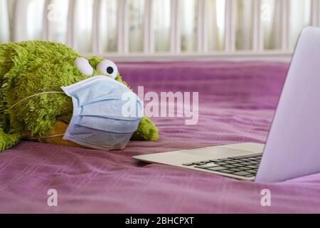 Green soft toy wearing medical face mask and looking at the computer screen on top of bed. Work at home. Stock Photo