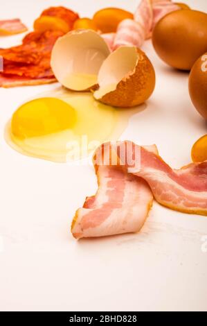 Broken chicken egg, scattered chicken eggs, slices of sausage and bacon and tomatoes on a white background. Close up Stock Photo