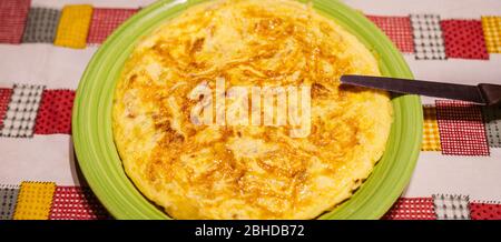 Tasty Spanish tortilla on a green plate with a knife to cut the portions on a table with a family tablecloth. Yellow omelette with eggs, potato and on Stock Photo