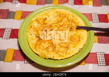 Tasty Spanish tortilla on a green plate with a knife to cut the portions on a table with a family tablecloth. Omelette with eggs, potato and onion. Stock Photo