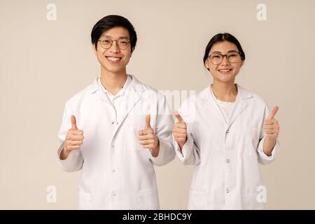 Image of asian young medical doctors in white uniform smiling and gesturing thumbs up isolated over beige background Stock Photo