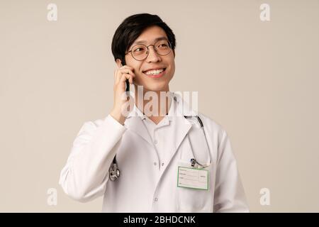 Image of asian young male doctor in medical white uniform smiling and talking on cellphone isolated over beige background Stock Photo