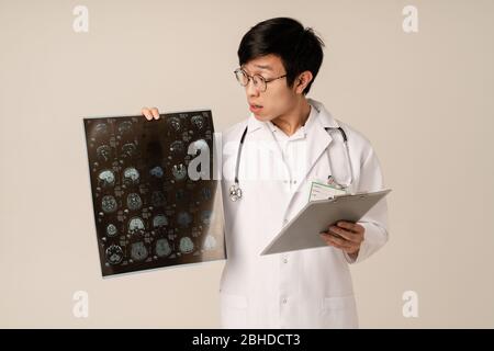 Image of asian young male doctor in uniform holding x-ray scan computed tomography results isolated over beige background Stock Photo
