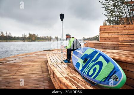 Athlete in wetsuit with paddle and surf sitting at wooden pier near the lake