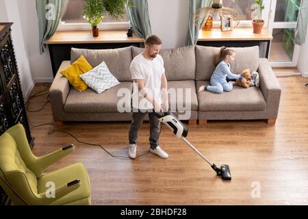 Young man cleaning floor of living-room with vacuum cleaner while his cute little daughter in pajamas playing with teddybear on couch near by Stock Photo
