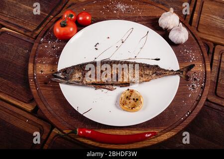 Appetizing mackerel fish baked on fire with spices, served on a plate and decorated with vegetables on wooden background Stock Photo