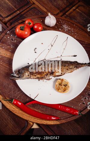 Appetizing mackerel fish baked on fire with spices, served on a plate and decorated with vegetables on wooden background Stock Photo