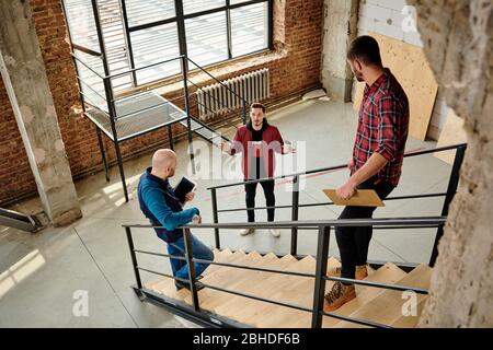 Two young engineers in casualwear standing on staircase inside unfinished building and listening to their colleague with glass of coffee Stock Photo
