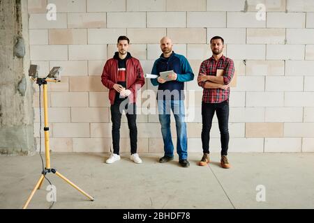 Three young male engineers or builders in workwear standing in line along wall of unfinished building in front of camera during work Stock Photo