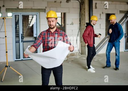 Young builder in hardhat and casualwear looking at sketch in blueprint on background of two colleagues talking at coffee break Stock Photo