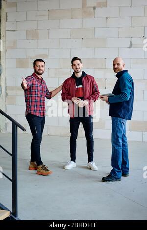Three young successful engineers or builders in casualwear standing inside unfinished building in front of camera and having start-up meeting Stock Photo