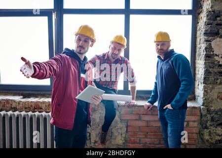 Young architect in helmet using touchpad while explaining his colleagues where to make wall at start-up meeting in unfinished construction Stock Photo