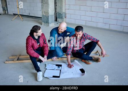 Happy young engineers sitting on the floor inside unfinished building and discussing points of sketch on blueprint at working meeting Stock Photo