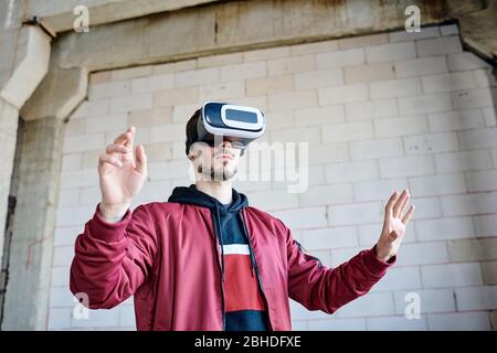Young engineer in vr goggles standing in front of virtual display and touching it while looking through examples of built constructions Stock Photo