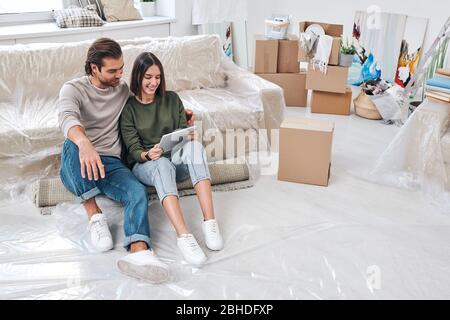 Pretty young woman with tablet and her affectionate husband looking through online furniture for their new house or flat in living-room Stock Photo