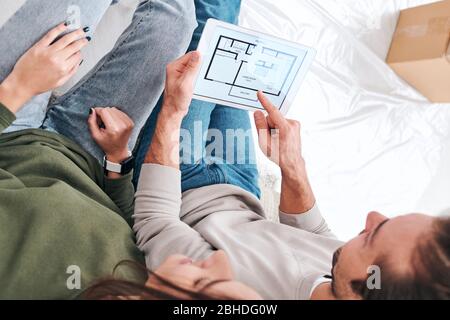 Young man pointing at tablet screen while having rest after removing and discussing sketch of new flat or house on the floor Stock Photo
