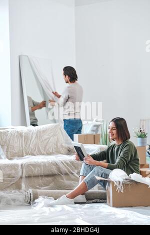 Happy young woman looking at picture in frame while sitting on the floor on background of her husband unpacking mirror in front of wall Stock Photo