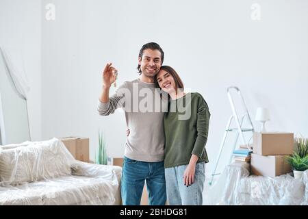 Young affectionate woman embracing her husband with key to their new house or flat while both standing in room with unpacked stuff Stock Photo