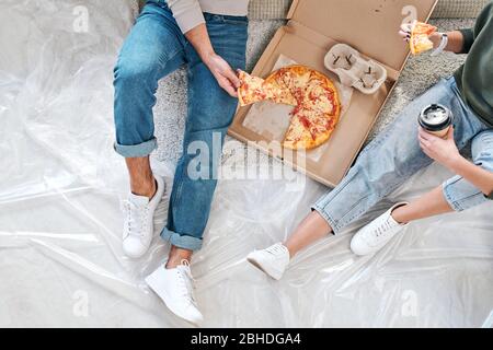 Overview of young couple in casualwear sitting on the floor and having pizza from box and coffee after removing to new flat or house Stock Photo