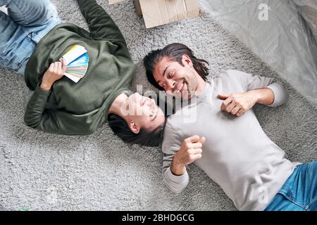 Top view of happy young amorous couple lying on the floor in their new house or flat after removing and discussing ideas Stock Photo