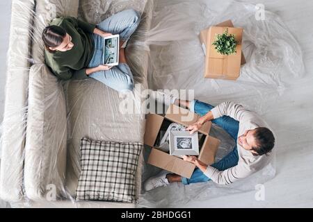 Top view of young woman with tablet and her husband with picture in frame sitting in front of each other and chatting in new flat Stock Photo