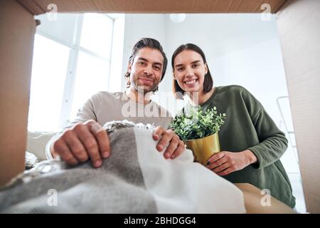 Young cheerful husband and wife in casualwear looking at you while unpacking boxes with household stuff in their new flat Stock Photo