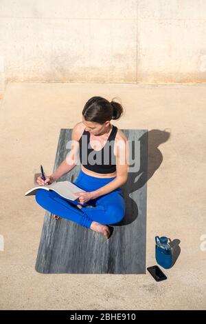 Sporty woman sitting on an exercise mat outside writing in a notebook, journaling. Stock Photo