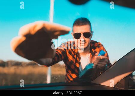 Young man carefully cleaning a windshield on his car with microfiber cloth and spray in a bottle. Stock Photo