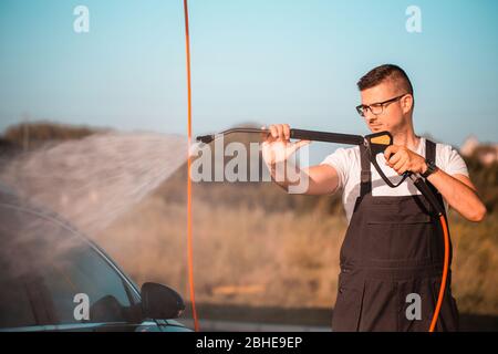 Young man washing car outdoors in a car wash station using high pressure water. Stock Photo