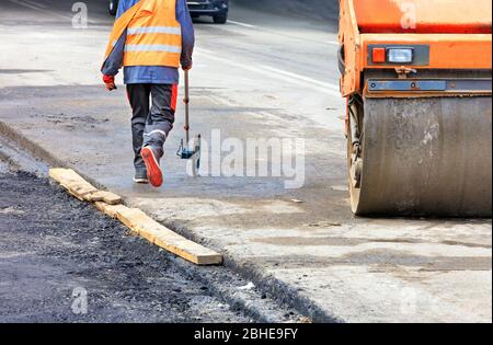 An engineer measuring road distances with a measuring wheel walks near a large vibratory roller. Stock Photo