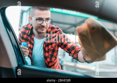 Young man carefully cleaning the windows on his car with microfiber cloth and spray in bottle. Stock Photo