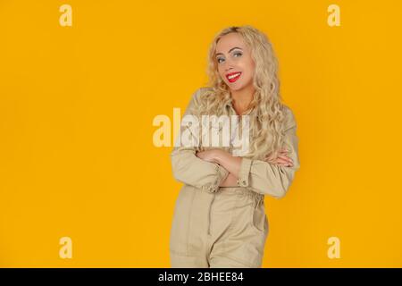 Portrait of a joyful and cheerful cute European curly-haired blonde woman who jokes cheerfully, entertains the conversation, smiles and laughs at the camera over the yellow wall. What a wonderful day Stock Photo