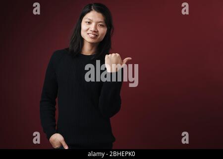 Image of joyful asian man smiling and pointing thumb aside isolated over burgundy background Stock Photo