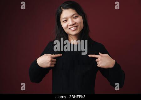 Portrait of a cheerful young asian woman standing isolated over dark background, wearing casual clothes, pointing fingers at herself Stock Photo