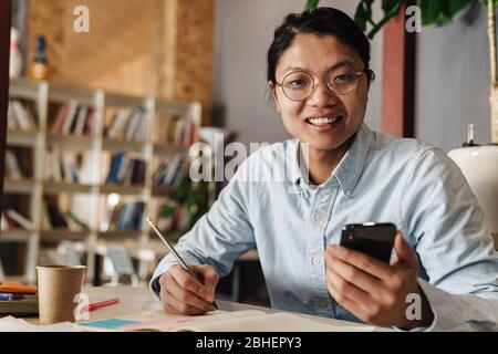 Image of smiling asian man in eyeglasses using cellphone and writing on exercise book while sitting at table Stock Photo