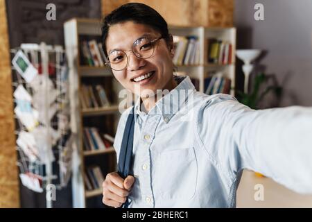 Image of cheerful asian man in eyeglasses smiling and taking selfie photo while standing at classroom Stock Photo