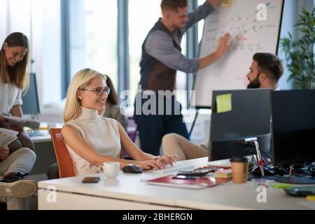 young business woman smiling and typing on a keyboard, with colleagues discussing plan on a whiteboard in the background. Dynamic business  space Stock Photo